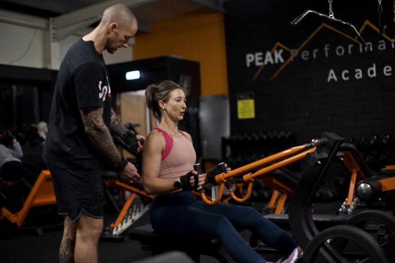 A woman is using a rowing machine in a gym under the guidance of a tattooed male instructor. The gym has various equipment and a "Peak Performance Academy" sign on the wall in the background. The lighting is focused on the subjects.