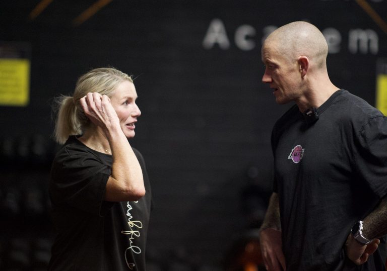 A woman and a man are standing and conversing in what appears to be an indoor gym. Both are wearing dark t-shirts. The woman has her hand up near her head, while the man has his hands by his sides. The background is dimly lit with blurred text visible.