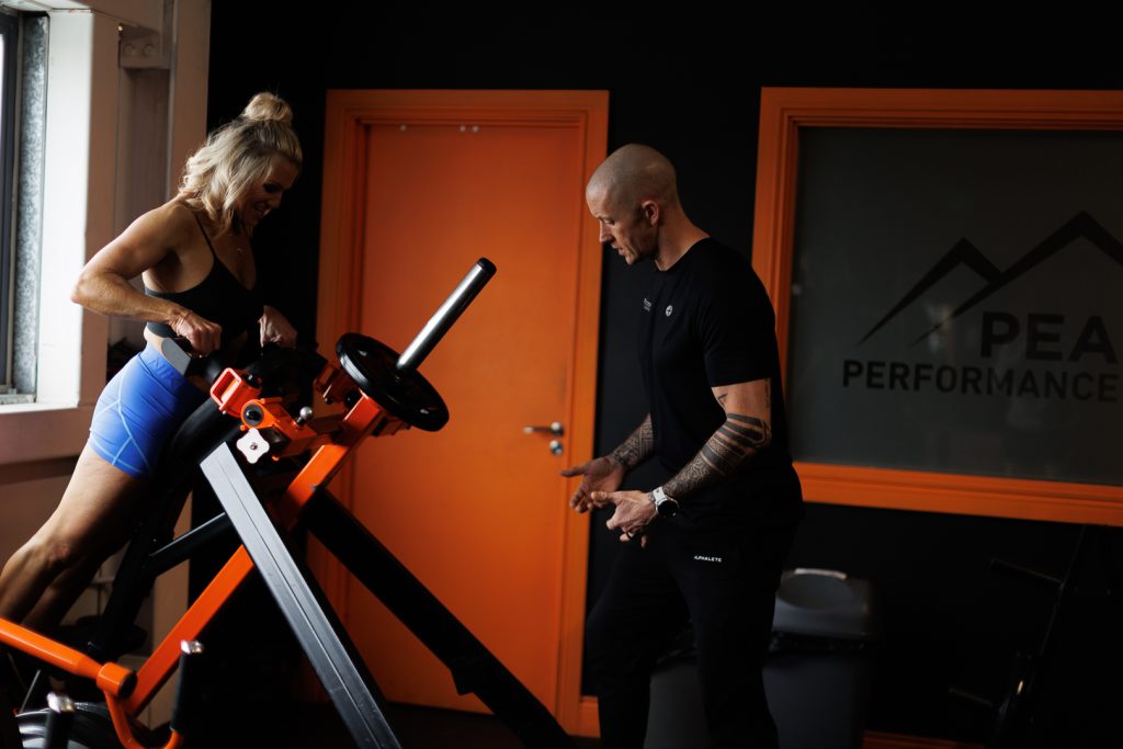 A woman in blue workout attire exercises on a fitness machine while a man in black workout gear, with tattoos on his left arm, supervises. They are in a gym with a window and a door, both with orange frames. A sign reading "Peak Performance" is partially visible.