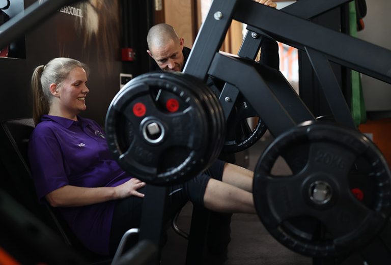 A woman is sitting on a leg press machine, wearing a purple shirt and black pants, while a man appears to instruct her. The leg press machine is loaded with weight plates. Both individuals are smiling.
