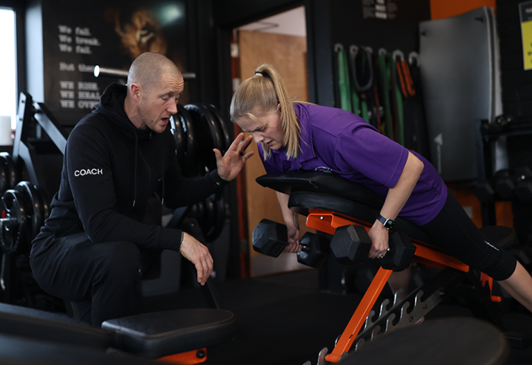 A woman in a purple shirt is performing dumbbell rows on an inclined bench in a gym. A man in a black hoodie labeled "Coach" is beside her, gesturing with his hand, presumably giving instructions or guidance. Various gym equipment is visible in the background.