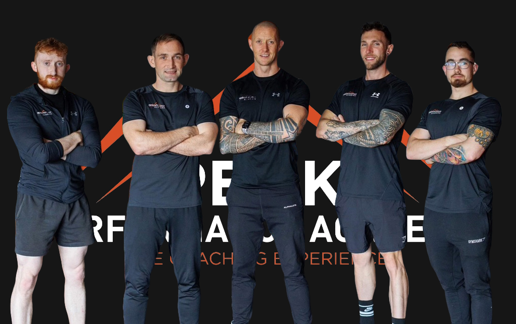 Five men wearing athletic wear stand side by side in front of a dark background with the text "Peak Performance Academy" partially visible behind them. Four of the men have their arms crossed, and two of them have visible tattoos on their arms.
