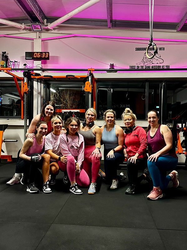 A group of nine women is gathered inside a gym with various exercise equipment in the background. They are dressed in athletic wear and are smiling, with some kneeling in the front and others standing behind. A neon-lit clock displays the time as 06:33.