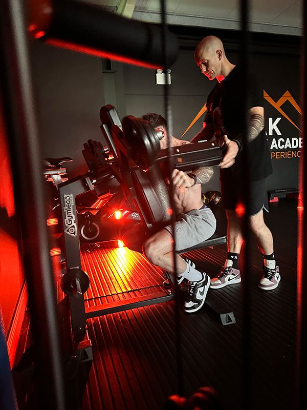 Two individuals in a gym: one person is performing a leg press exercise on a machine, while the other is supervising. The gym has dark walls and is lit by red lights. Gym equipment and a large logo can be seen in the background.